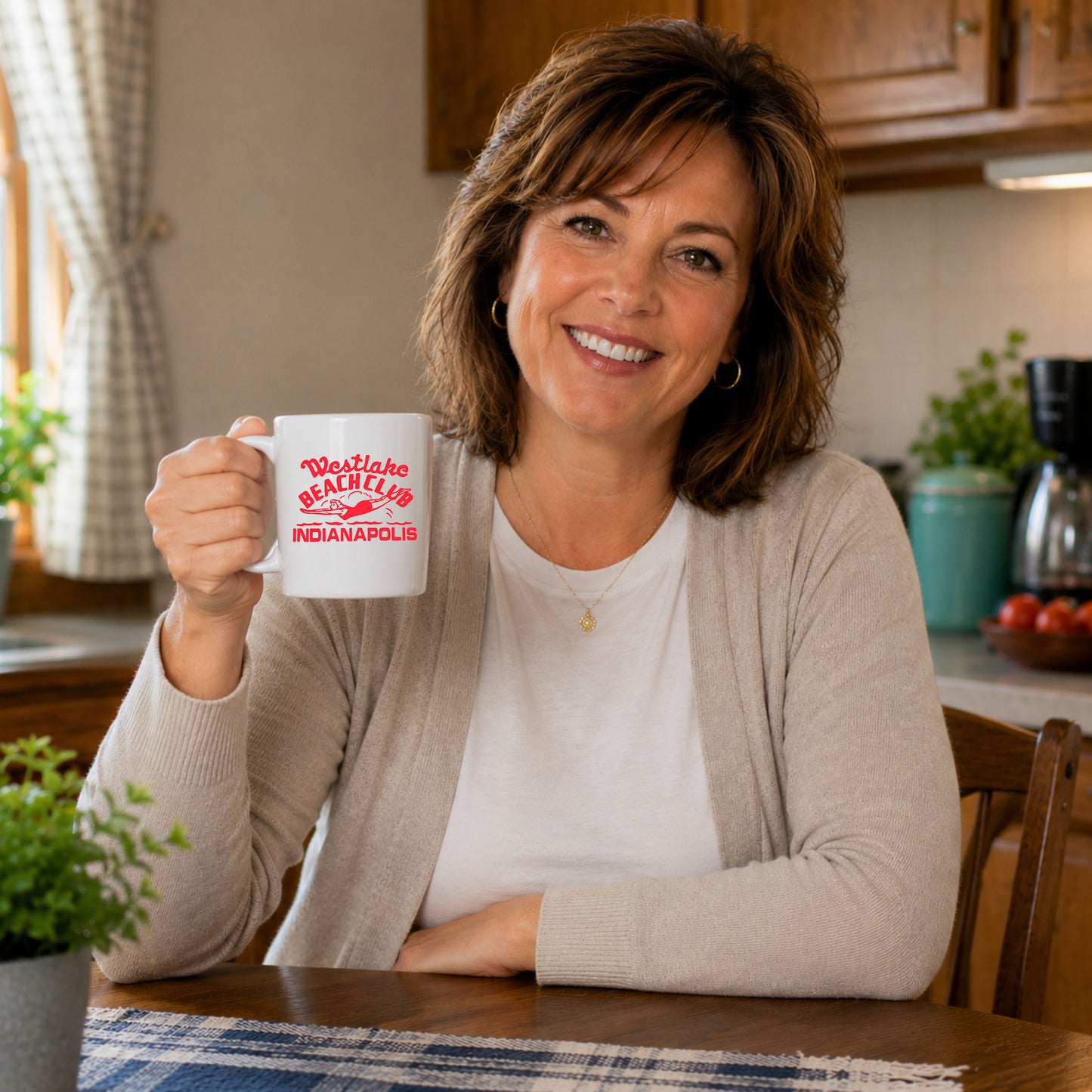 Woman holding white Westlake Beach Club Indianapolis nostalgia coffee mug while sitting at a kitchen table in a cozy home kitchen