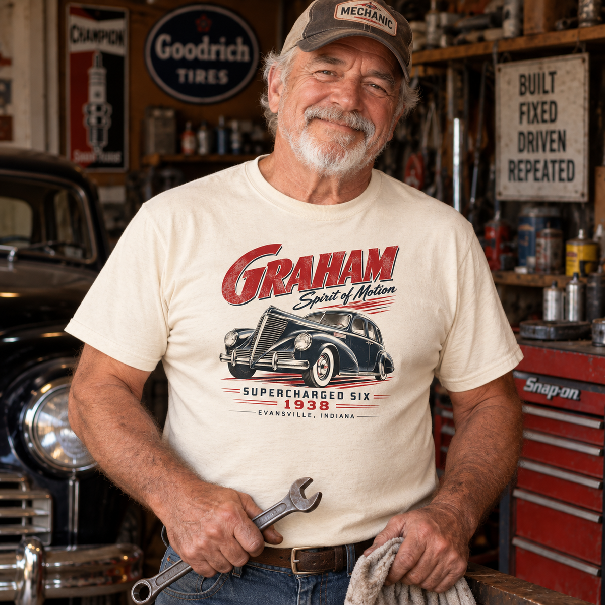 Older man wearing a natural Gildan 5000 T-shirt featuring a vintage 1938 Graham Supercharged Six Evansville Indiana automobile design in a garage.