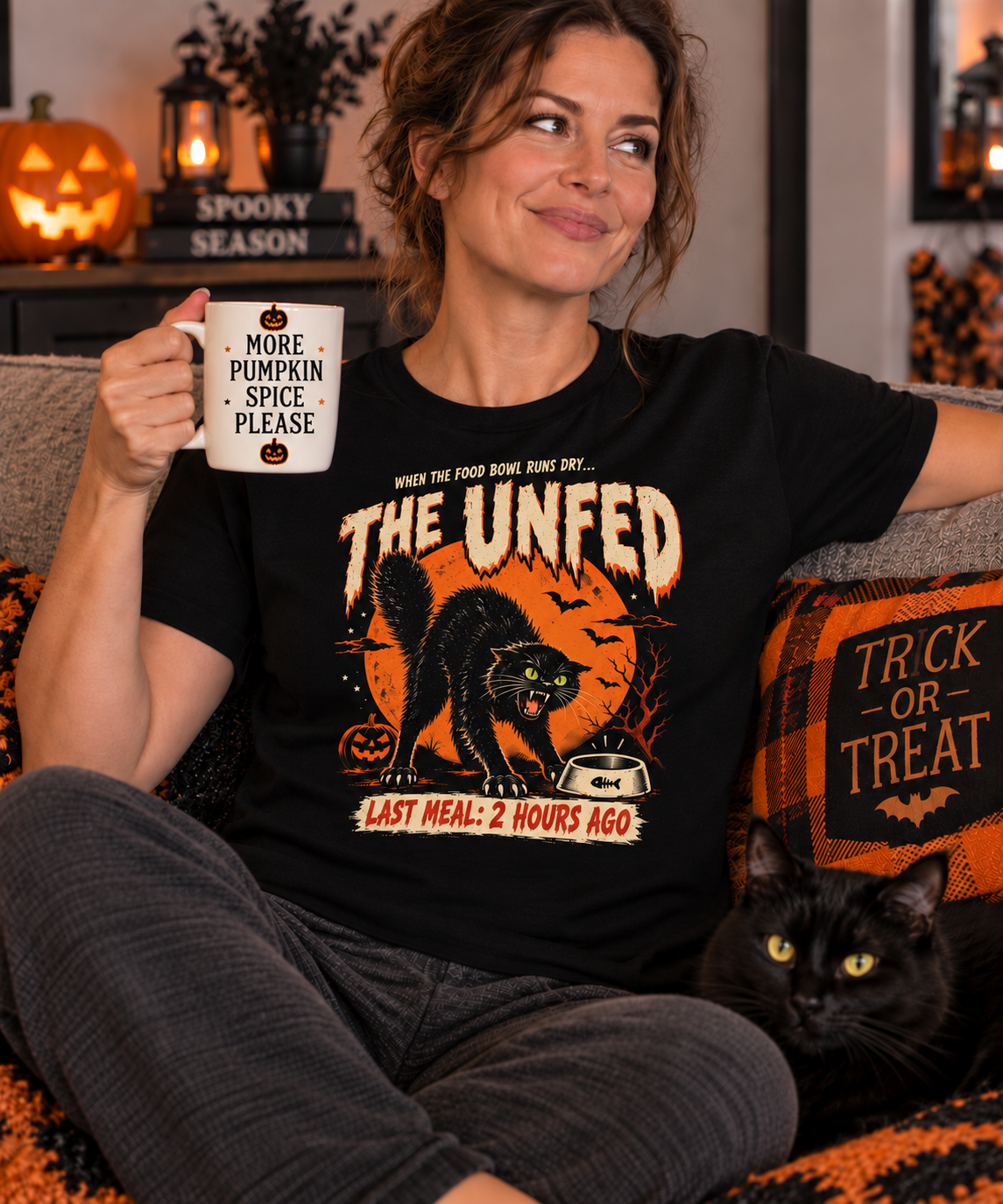 Woman wearing a black Gildan 5000 T-shirt featuring The Unfed funny black cat Halloween design while sitting with a black cat and Halloween decorations.