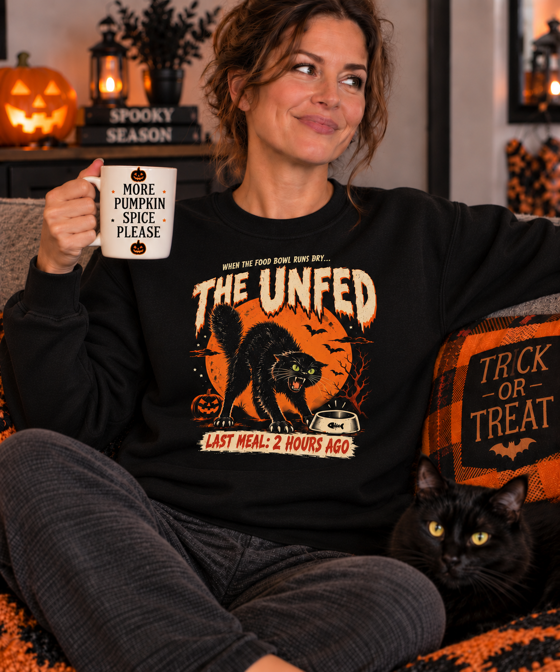Woman wearing a black Gildan 18000 sweatshirt featuring The Unfed black cat Halloween design while relaxing with a black cat and Halloween decorations.
